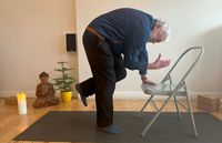 Ein älterer Mann in Freizeitkleidung übt mit Hilfe eines grauen Klappstuhls auf einer schwarzen Yogamatte. Der Raum ist schlicht und einladend gestaltet, mit einem hölzernen Dielenboden, einem hellen Wandfarbton und einer ruhigen Ecke.