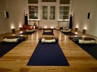 Ein Yoga-Raum bei sanfter Abendbeleuchtung, vorbereitet für eine Klasse, mit sechs dunkelblauen Matten auf einem hellen Holzboden. Jede Matte ist mit einem Yogakissen, einem Bolster und einer gefalteten Decke ausgestattet.
